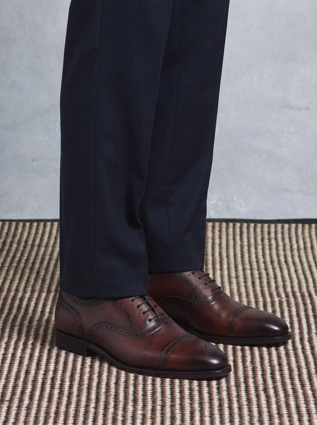 A close-up of a person's legs wearing dark navy trousers and brown wingtip oxford shoes with brogue detailing, standing on a woven rug against a textured gray wall.